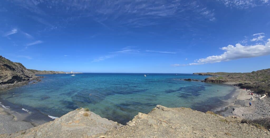 En la foto se aprecian 3 de las calan de la costa de cala Presili, en donde las aguas tranquilas de color turquesa contrastan con el azul claro del cielo al fondo se aprecia el Faro de Favaritx con colores blanco y azul oscuro.