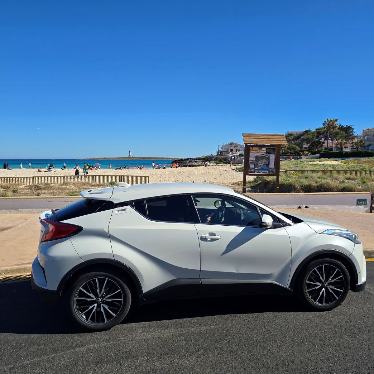 Preparar viaje a Menorca: Coche de alquiler junto a una playa de Menorca con el mar azul y un faro al fondo